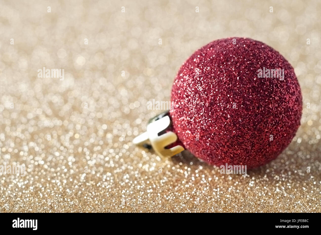 A single, sparkly red bauble, coated in glitter, resting on a gold glitter background that softens into soft focus bokeh in the background.  Copy spac Stock Photo