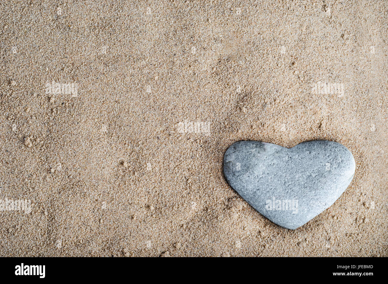 A grey heart shaped stone nestled in golden sand at bottom right of frame.  Copy space in sand. Stock Photo