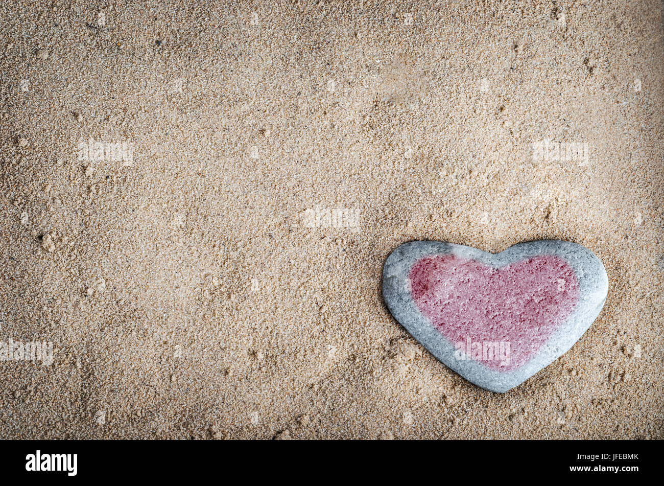 A grey heart shaped stone on grainy sand, tinted with a red heart.  This version is vignetted and edited to give a retro or lo-fi appearance. Stock Photo