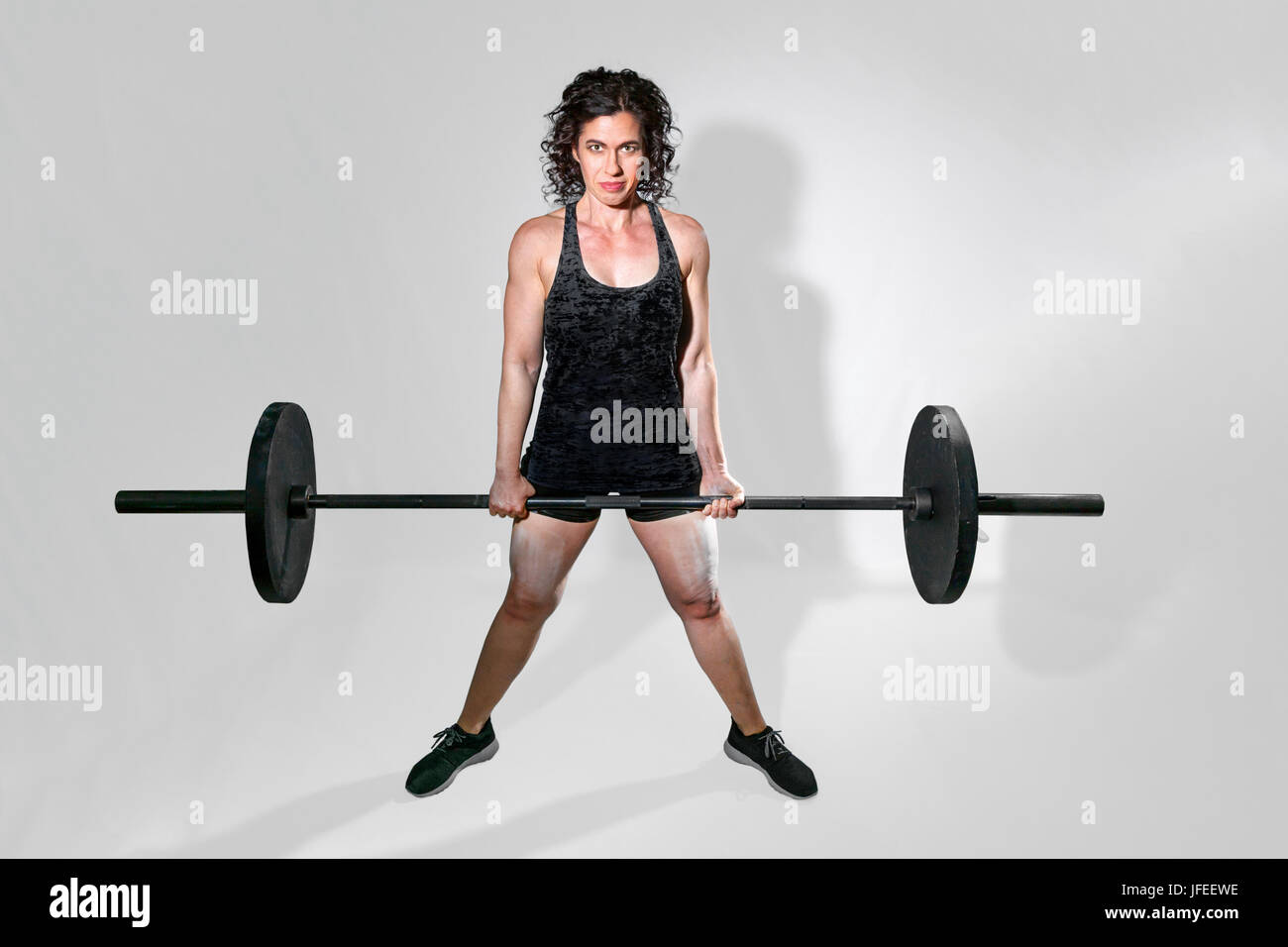 Tough, female weightlifter holds a barbell in a dead lift with an intense glare for the camera. There is a path around the woman. Stock Photo