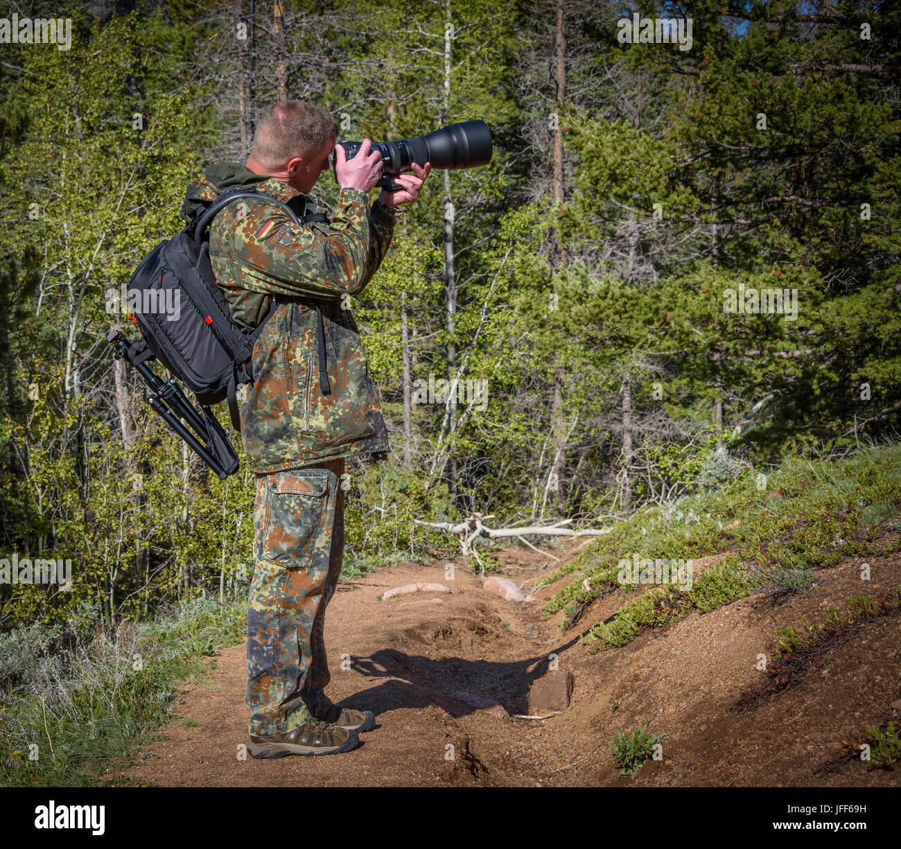 Wildlife, nature man photographer in camouflage outfit with a backpack and tripod standing on a mountain forest trail and shooting, taking pictures Stock Photo