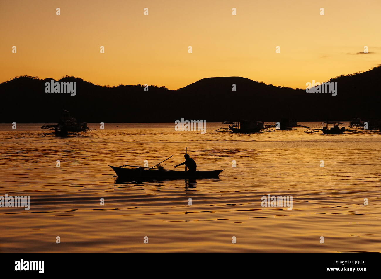 Tranquil sunset behind hills background. Lone fisherman paddles small boat setting out fishing net.  Golden sky reflecting in sea. Coron, Philippines. Stock Photo