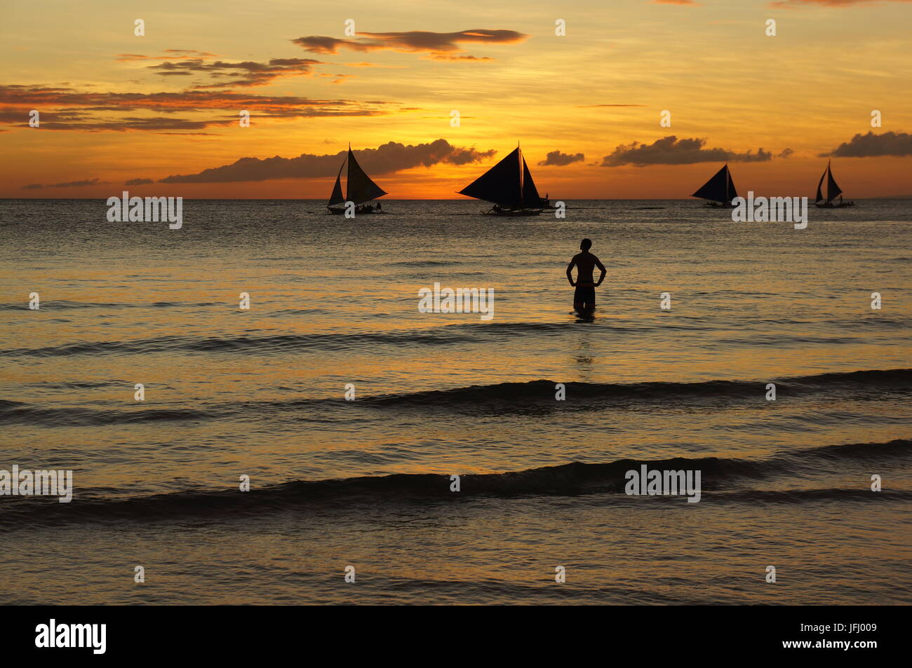 Beach sunset background stunning Philippines island Boracay.  A man stands hands on hips in the sea watching the sailing boats pass by the golden sky. Stock Photo