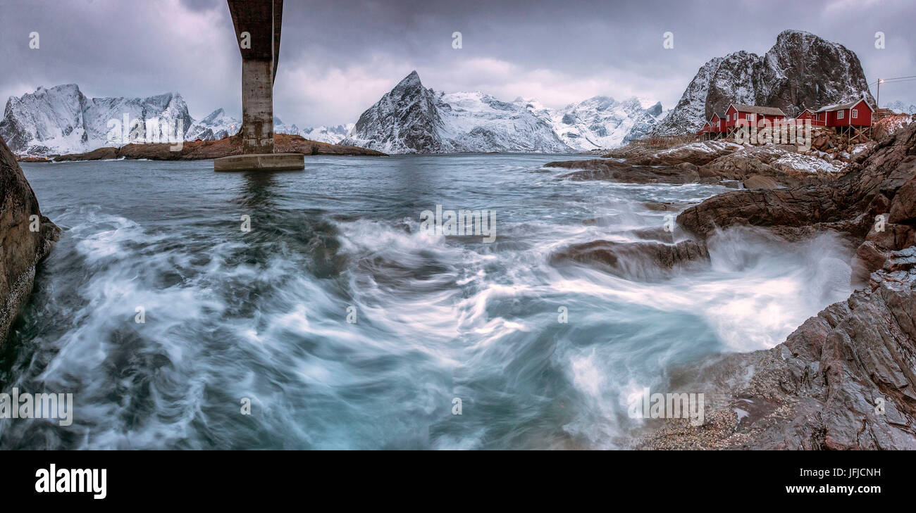 A bridge spans the rough sea and the cliffs with the houses of fishermen, Hamony, Lofoten Islands, Northern Norway, Europe Stock Photo