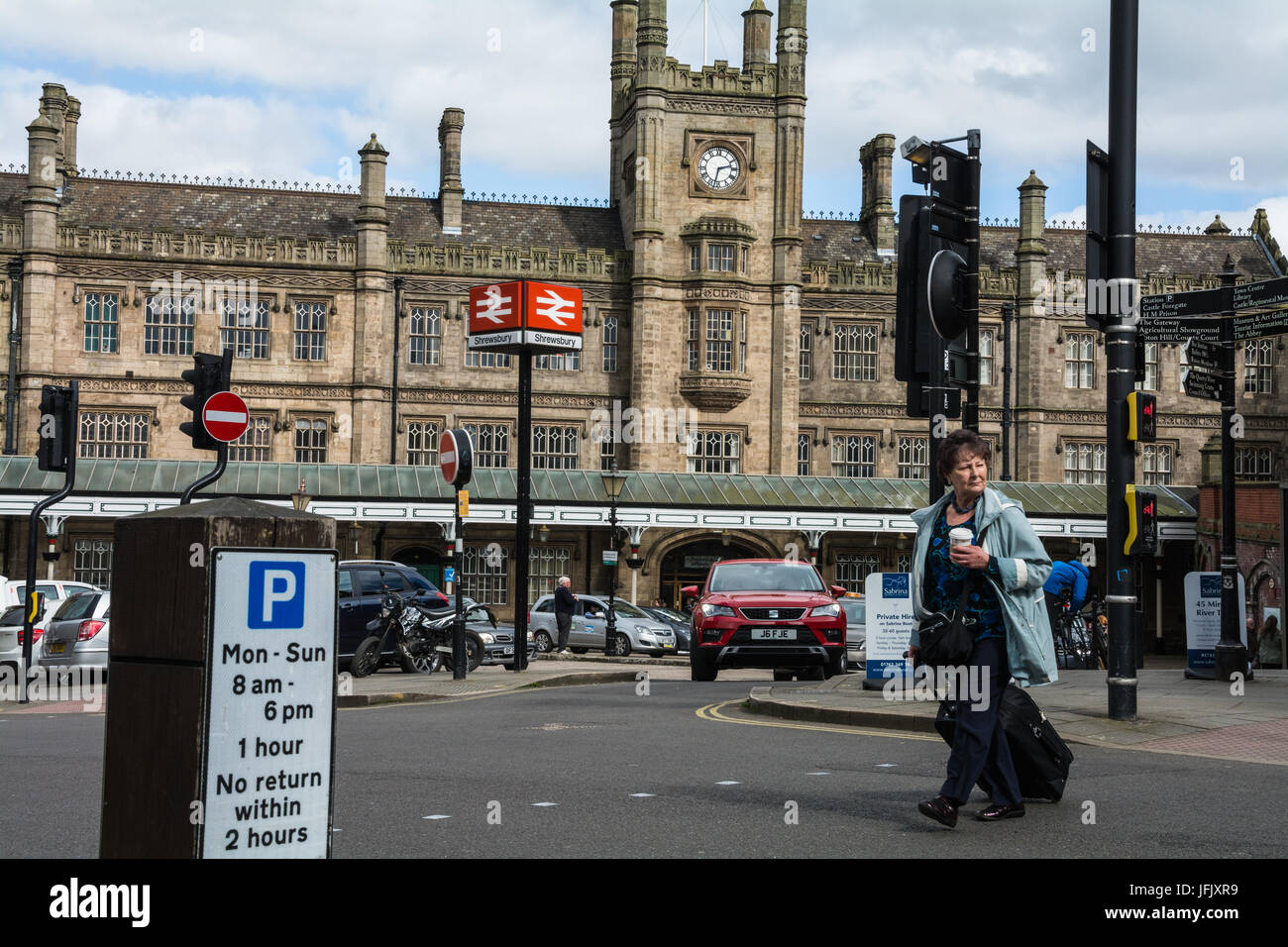Shrewsbury train station in the background. Commuter holding a cup of coffee, pulling a suitcase, crossing the A5191 road in the foreground. UK. Stock Photo