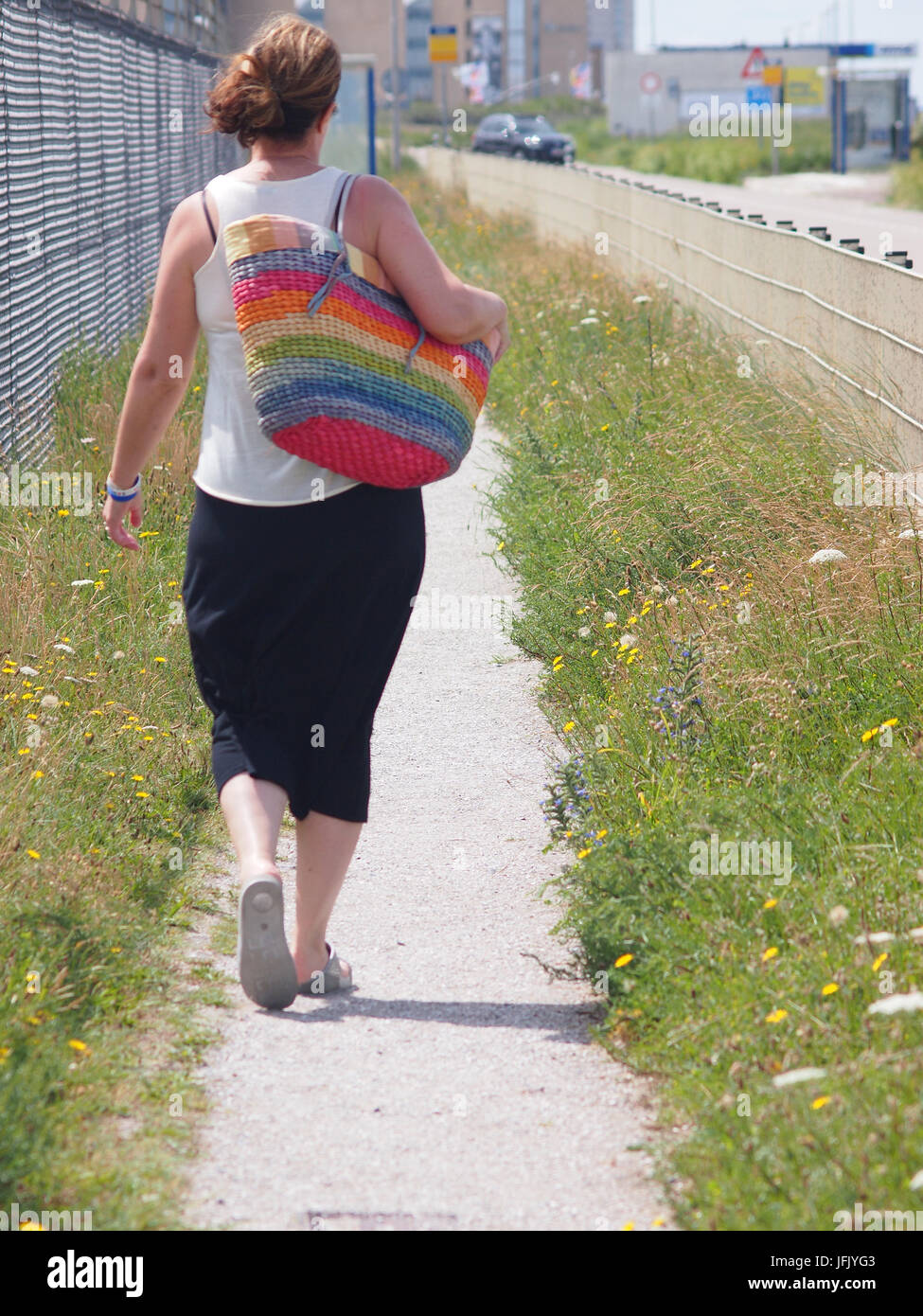 Woman wearing a black skirt and white top walking along a coastal path with a large stripey, colourful beach bag (seen from behind). Stock Photo