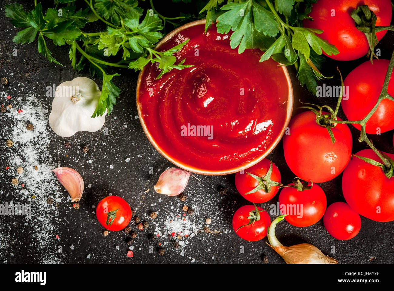 Fresh homemade organic tomato sauce or ketchup, in a small bowl. With the ingredients - parsley, onions, garlic, tomatoes, salt, pepper. On a black st Stock Photo