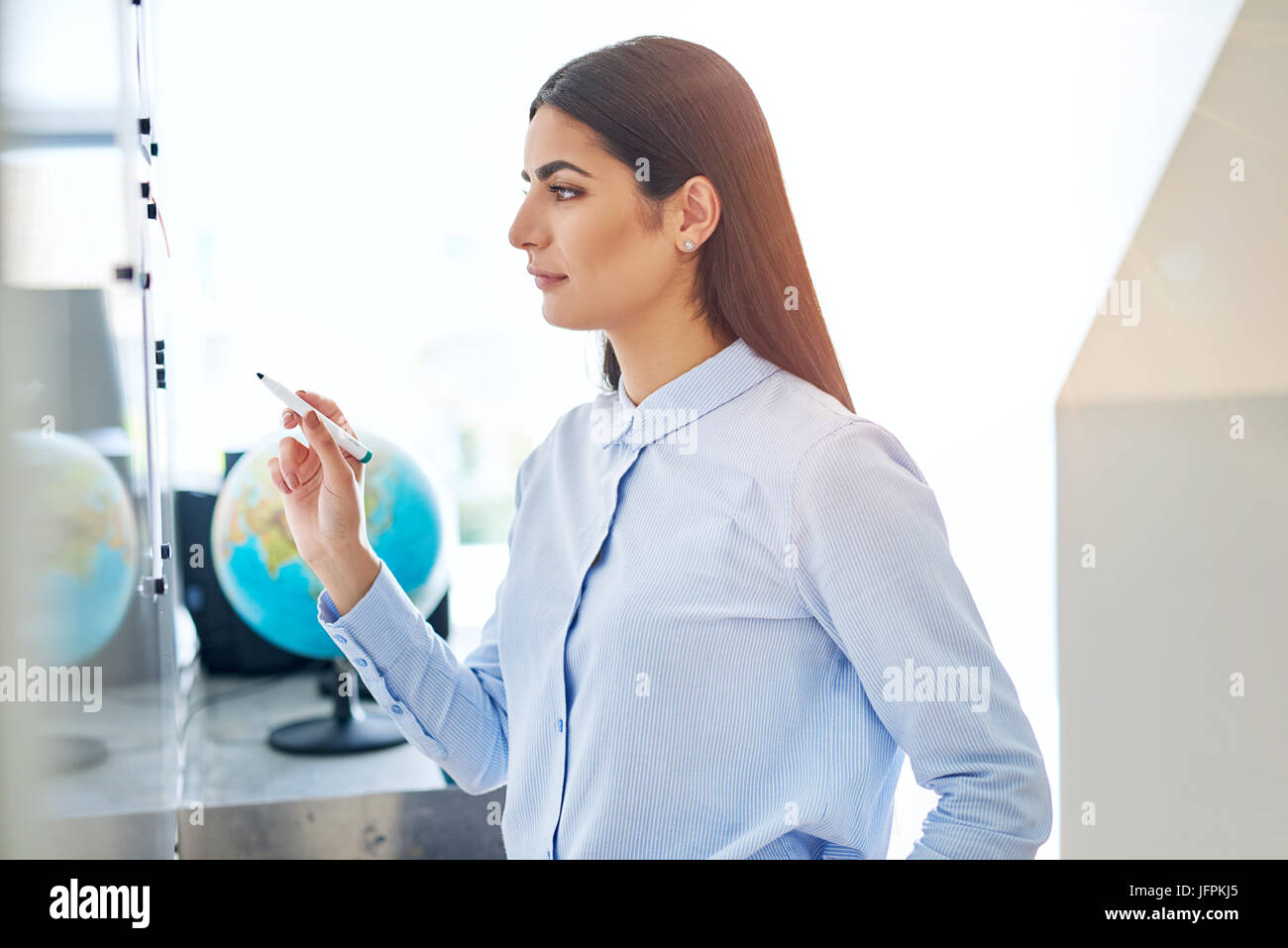 Serious young woman in blue at white board in classroom or small office with globe in background Stock Photo