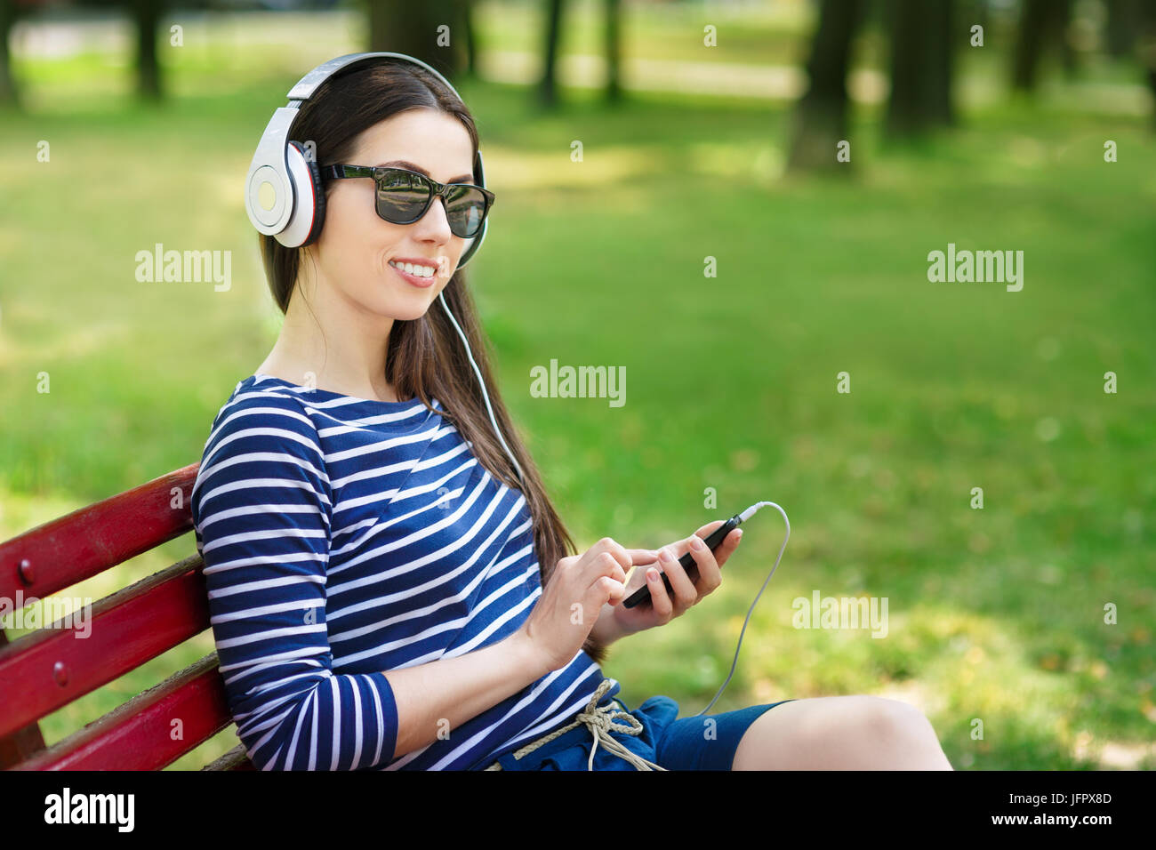 Young happy smiling brunette woman with headphones outdoors on summer day. Girl listening music in headphones in park Stock Photo