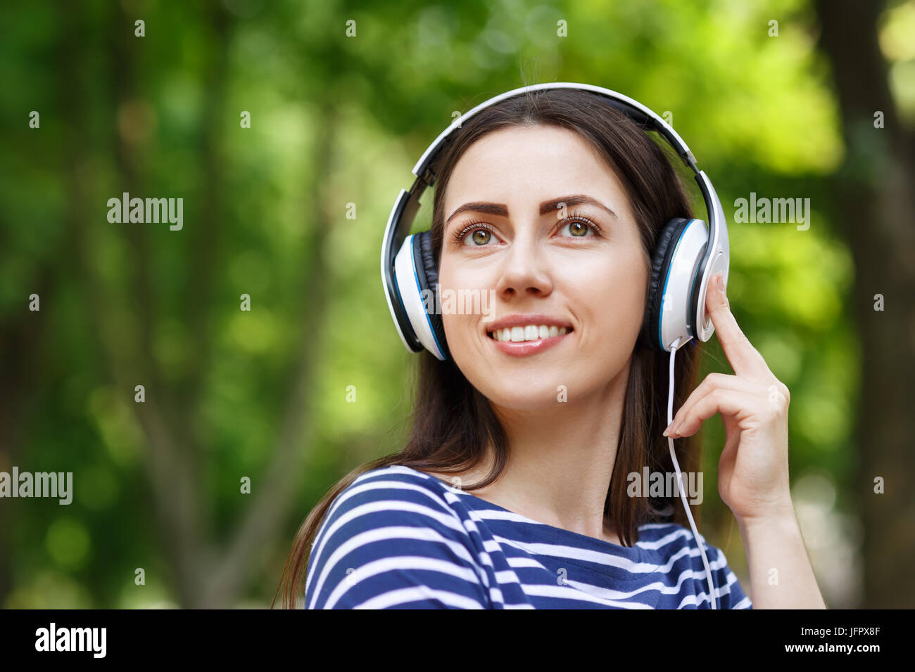 young happy smiling brunette woman with headphones outdoors on summer day. Girl listening music in headphones in park. Portrait of woman at outdoor wi Stock Photo