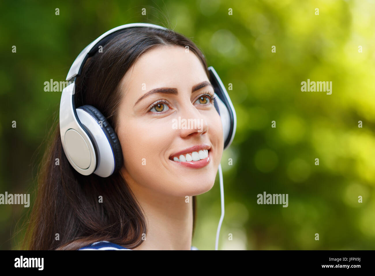 closeup portrait of young happy smiling brunette woman with headphones outdoors on summer day. Girl listening music in headphones in park Stock Photo