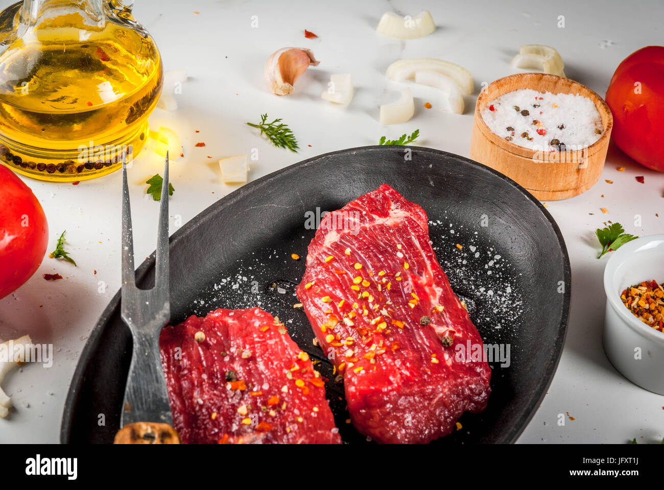 Meat, beef. Fresh raw steaks in a frying pan. Spices (salt, pepper), fresh vegetables - tomatoes, carrots, garlic, onions. On a white marble table, wi Stock Photo