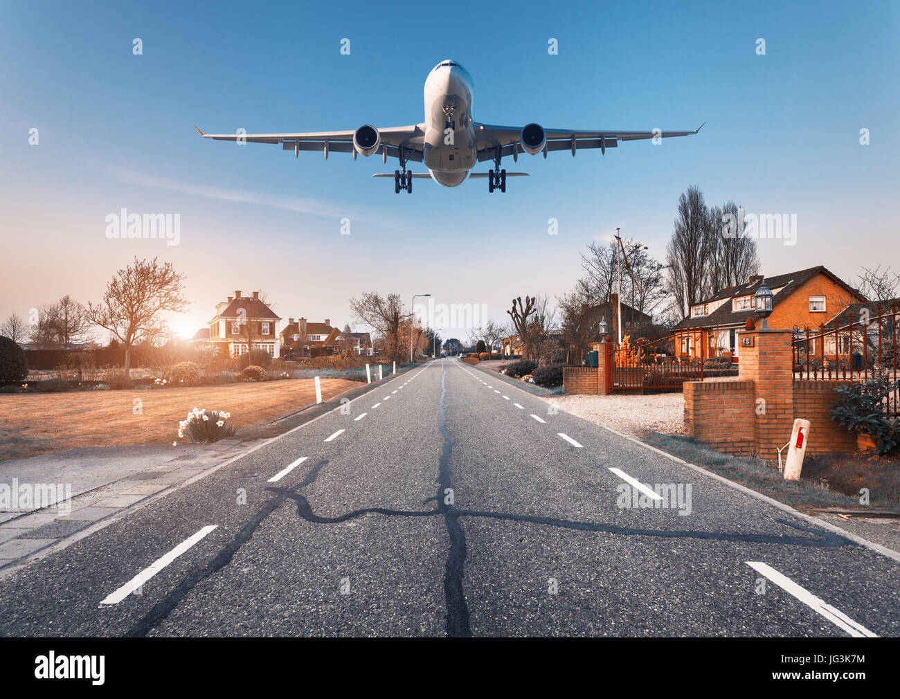 Beautiful cityscape with passenger airplane is flying in the sunset sky above the asphalt road  through the town with houses and courtyards at sunset  Stock Photo