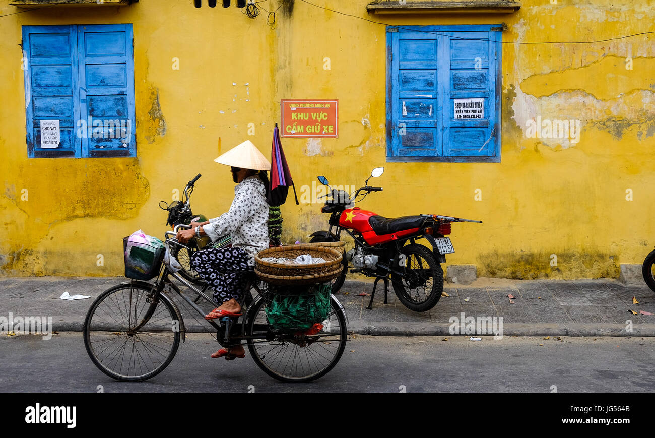 Vietnamese street vendor riding her bike along a street in Hoi An, Vietnam Stock Photo