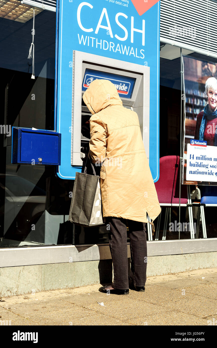 Elderly woman with a hooded coat uses an ATM Cash Point machine of a ...