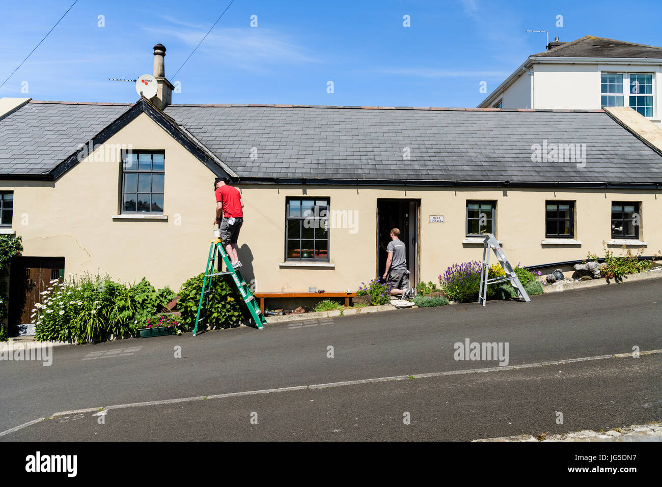 Two men painters painting a picturesque cottage on a steep hill road. Stock Photo