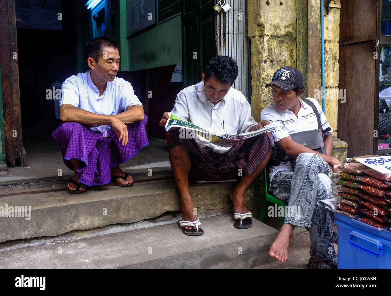 Three men share a newspaper whilst sitting outside a building in downtown Yangon, Myanmar Stock Photo