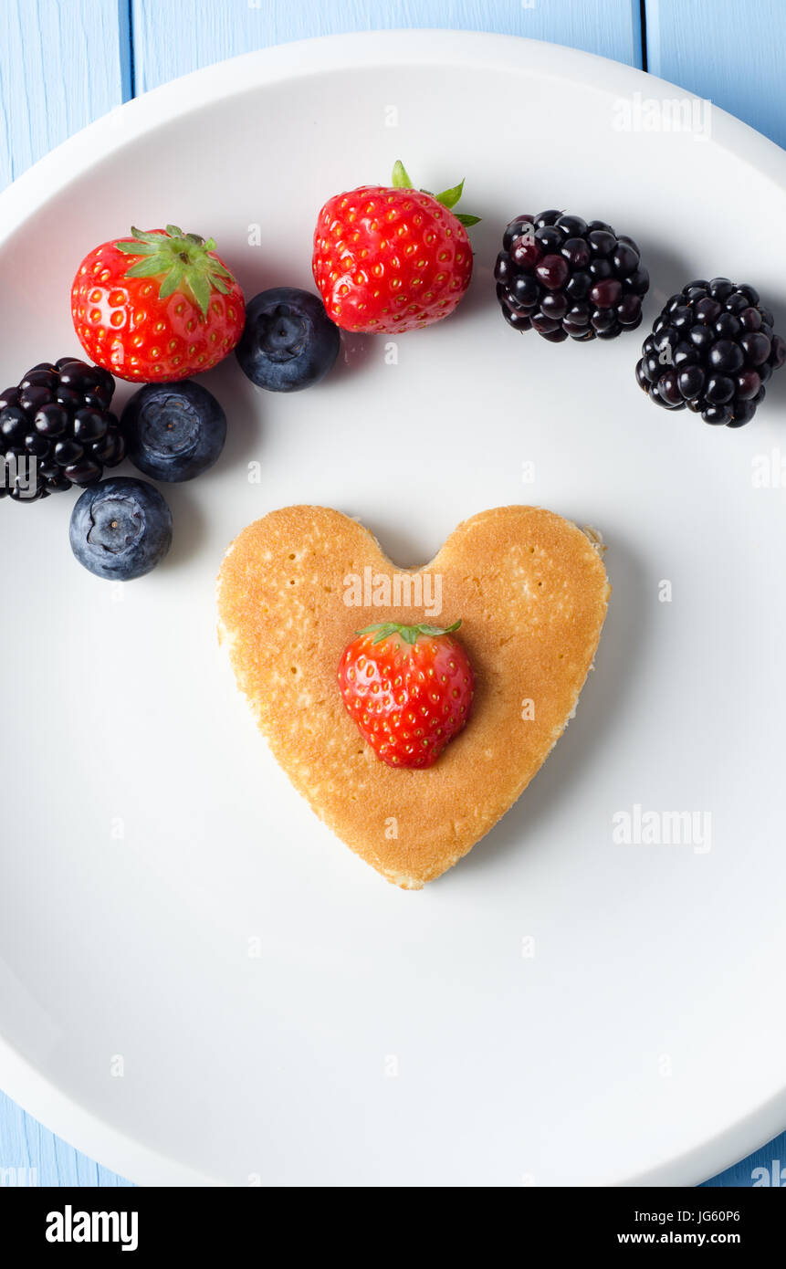 A heart shaped pancake in the middle of a white plate, with a strawberry centred on top of it.  Above it, a variety of Summer fruits are arranged in a Stock Photo