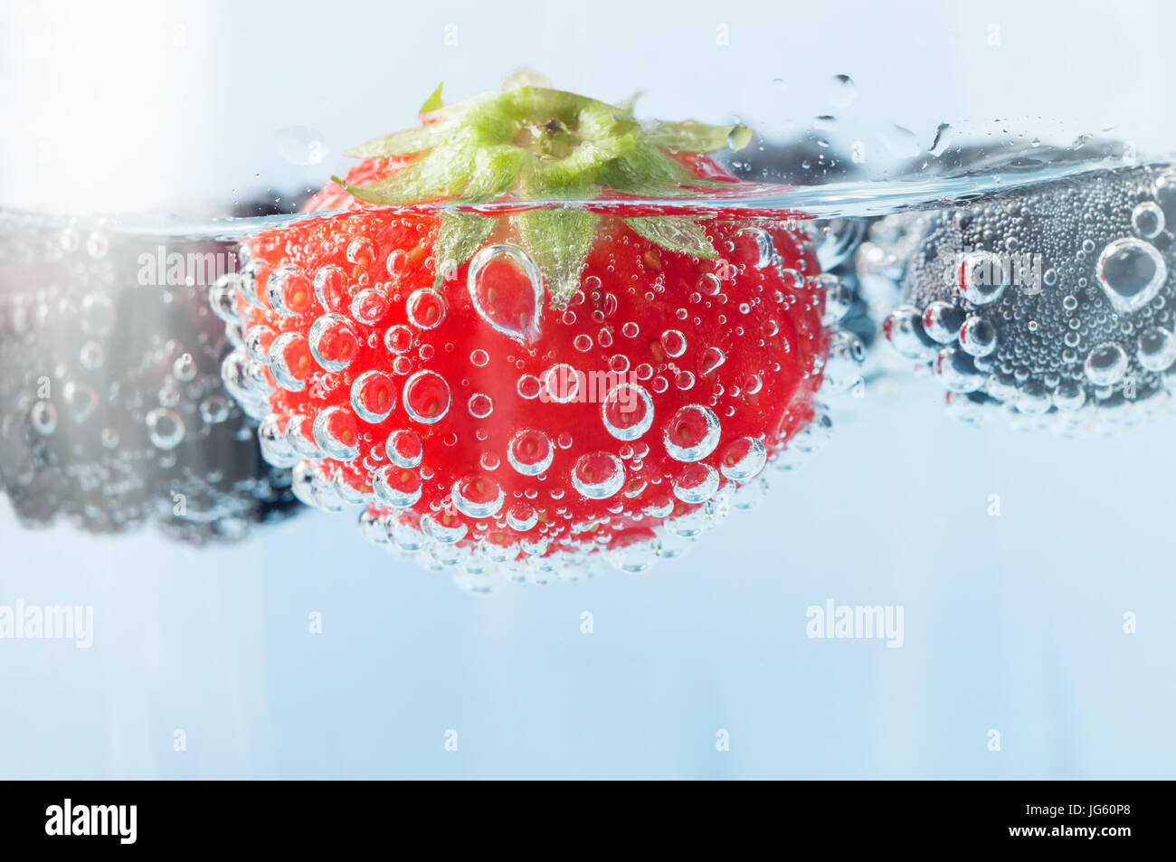 Close up (macro) of a bright red fresh strawberry, floating in sparkling water and covered in oxygen bubbles.  Blackberry and blueberries in soft focu Stock Photo