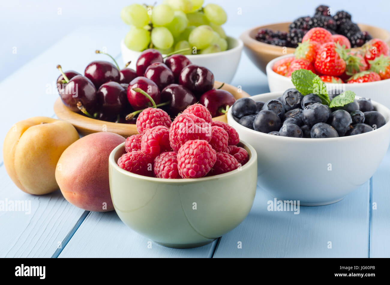 A selection of different Summer fruits, in a variety of bowls on a painted blue wood planked farmhouse kitchen table, against a light blue background. Stock Photo