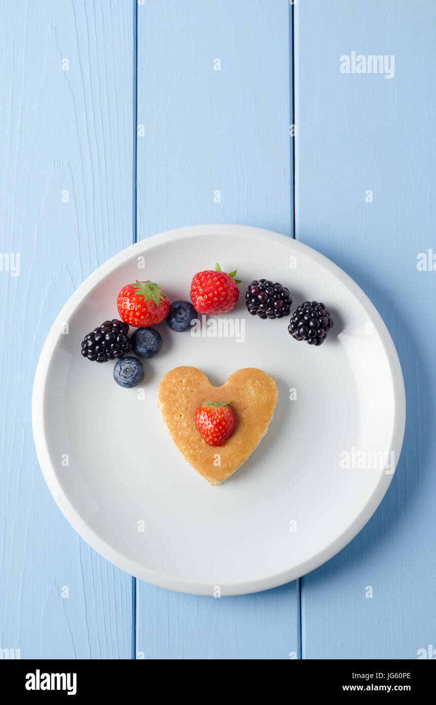 Overhead shot of a heart shaped breakfast pancake underneath an arc of Summer fruits and topped with a cut strawberry.  Food set on a white china plat Stock Photo