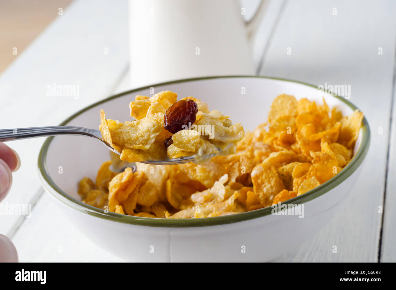A hand lifting a spoonful of cornflakes and raisins from a white bowl with green rim, on a painted white wood planked table with milk jug in soft focu Stock Photo
