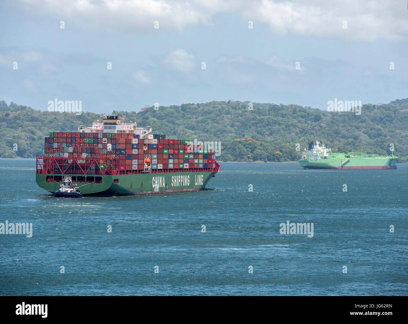 A Large Container Ship Xin Mei Zhou Of The China Shipping Line Passing Through Gatun Lake Of The Panama Canal, Steered By A Tug Boat Stock Photo