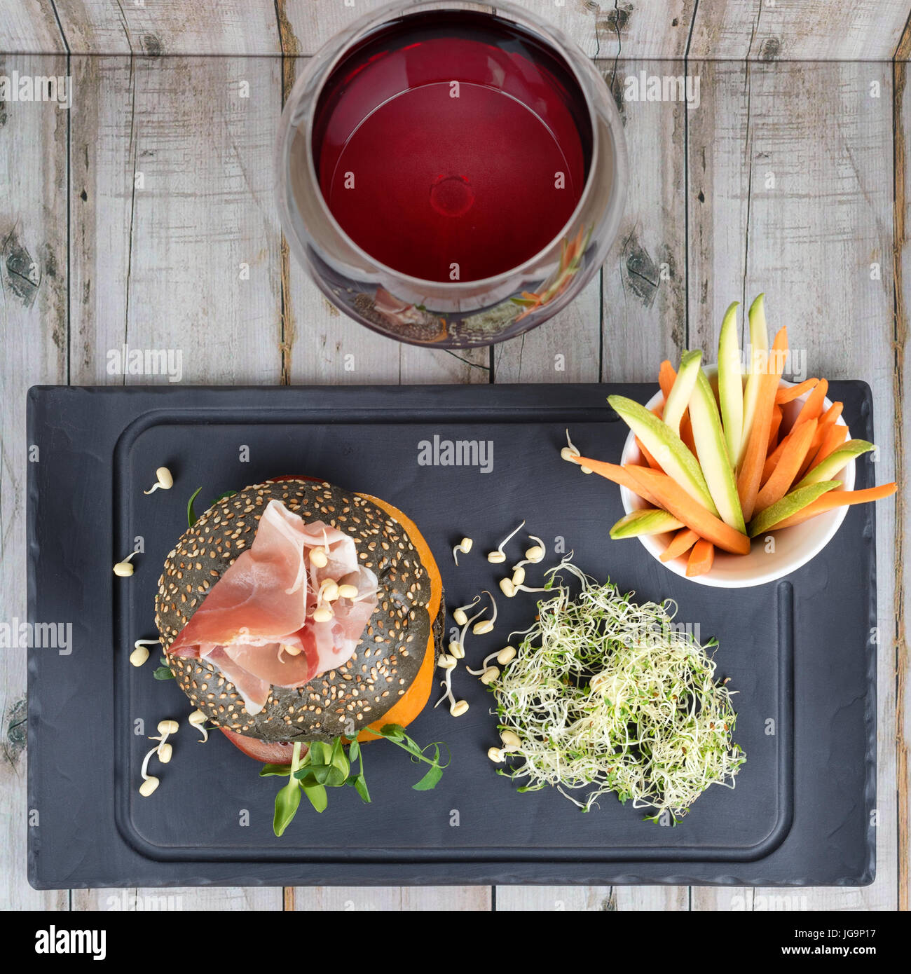 Healthy burger with hamon, tomatoes, micro greens and black wholegrain buns, vegetable sticks and red wine on black slate board over wooden background. Clean eating, dieting, detox food concept. Stock Photo