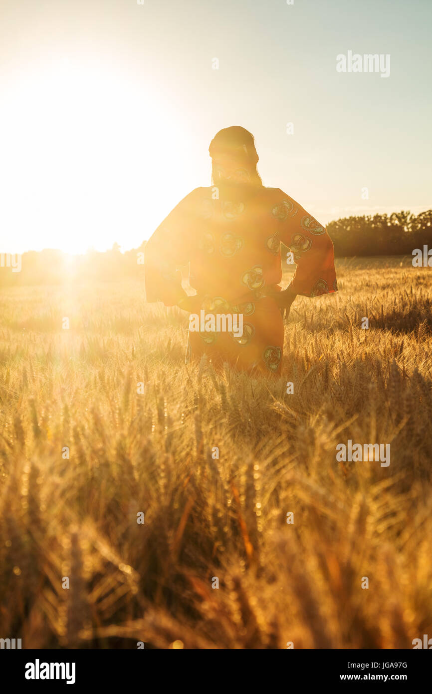 African woman in traditional clothes standing with her hands on her hips in field of barley or wheat crops at sunset or sunrise Stock Photo