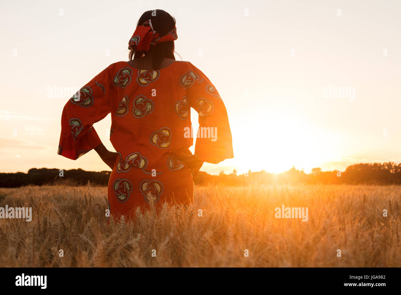 African woman in traditional clothes standing with her hands on her hips in field of barley or wheat crops at sunset or sunrise Stock Photo
