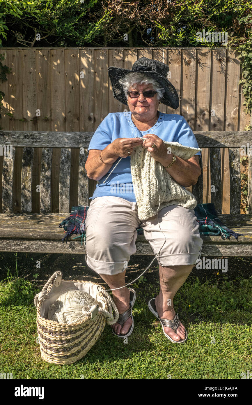 A smiling old / elderly / senior woman / lady knitting while seated on a wooden bench, alone in a garden in the warm sunshine. England, UK. Stock Photo