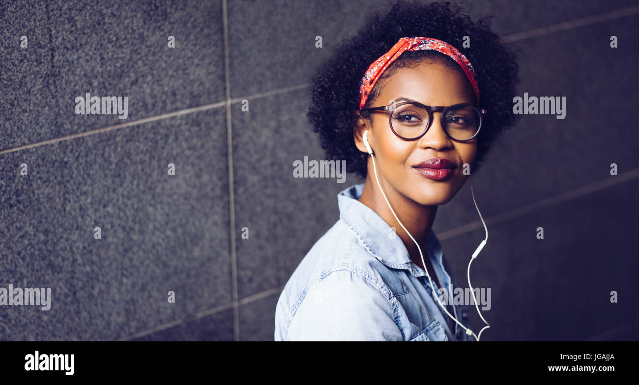 Confident young African woman wearing glasses and a bandana deep in thought while listening to music on earphones Stock Photo