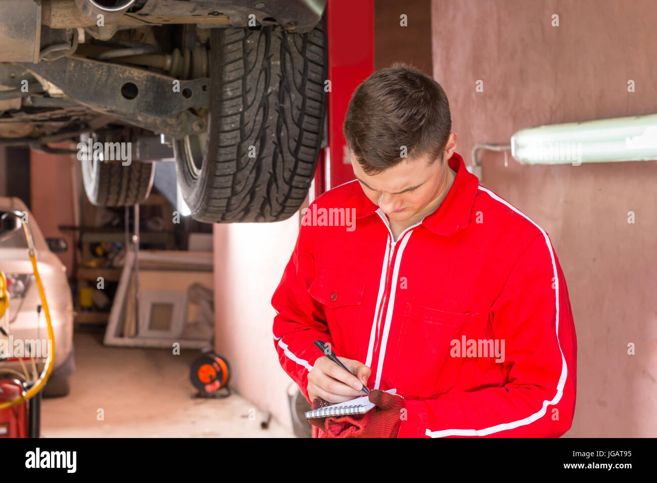 Young male auto mechanic standing making notes underneath a lifted car ...