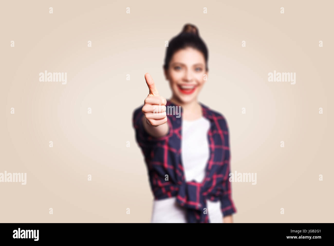 Young happy girl with casual style and bun hair thumbs up her finger, on beige blank wall with copy space looking at camera with toothy smile. focus o Stock Photo