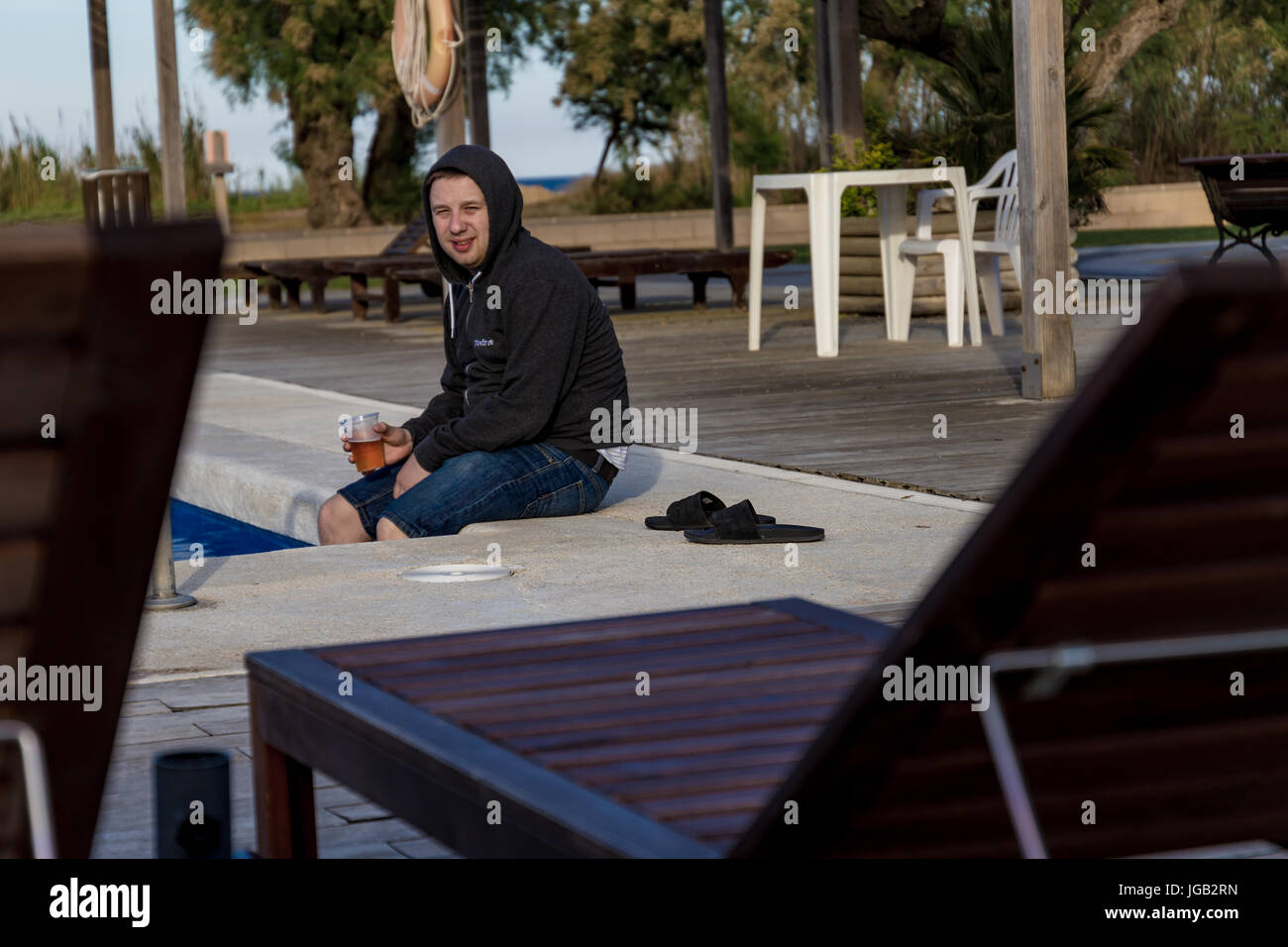 Student in a hoodie drinking beer and tipping legs into pool water Stock Photo