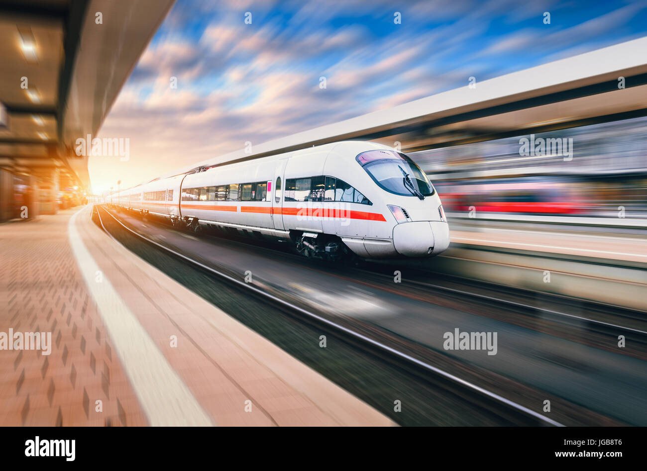 White modern high speed train in motion on railway station at sunset. Train on railroad track with motion blur effect in Europe in evening. Railway pl Stock Photo