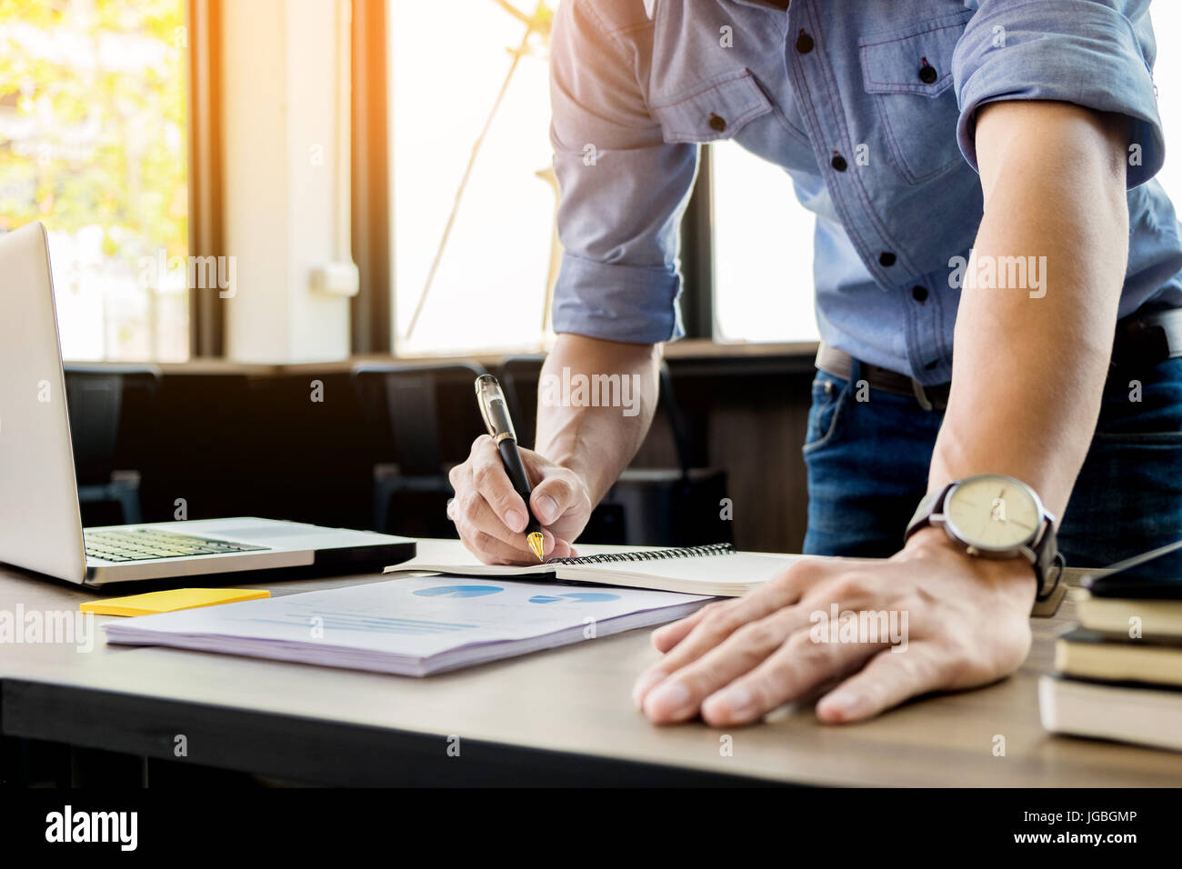 Young business man working in bright office, using laptop, writing notes. Stock Photo