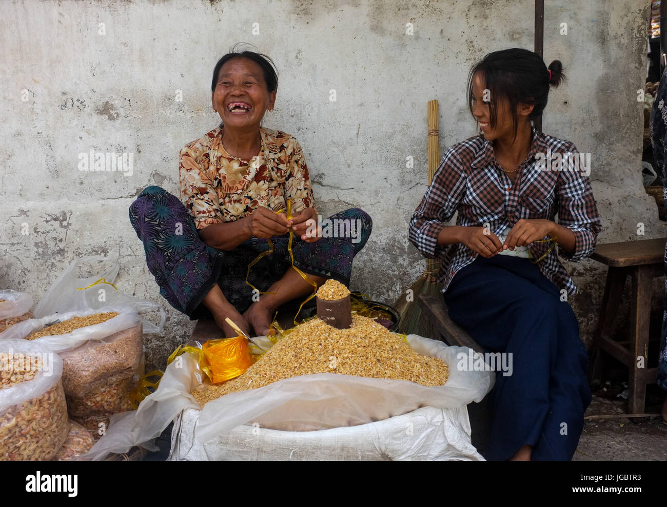 Two Burmese women laughing whilst sitting at their market stall in Mandalay, Myanmar Stock Photo