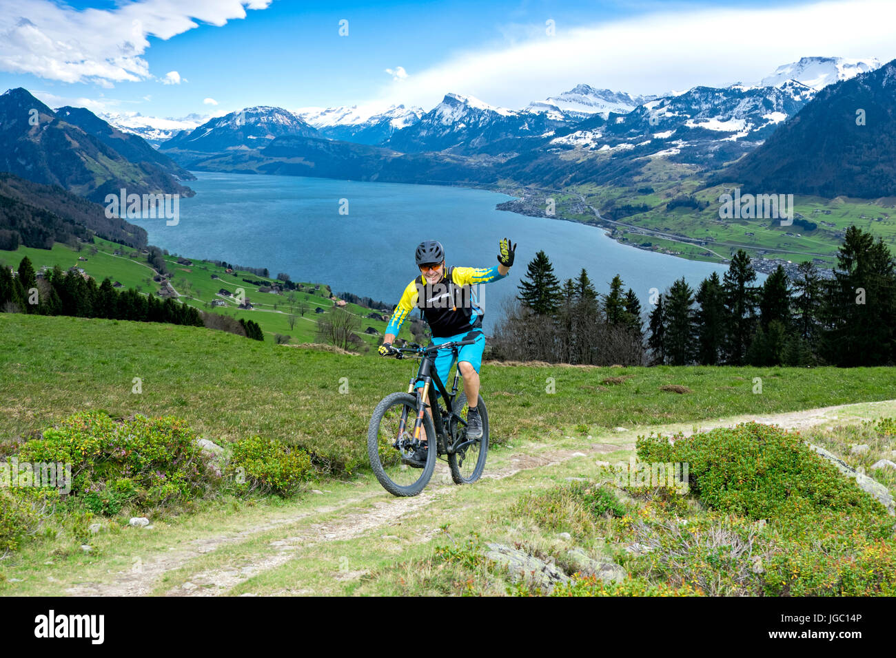 Sportive man in middle age with mountain bike on mountain trail beckons the viewer. The background shows the Lake Lucerne. Stock Photo