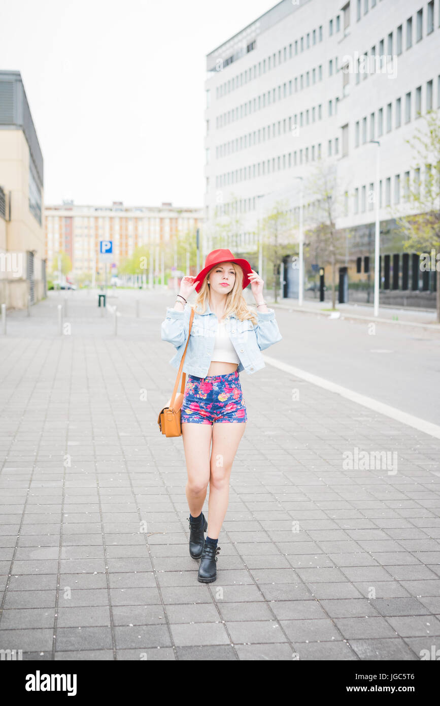Young beautiful blonde caucasian girl walking in the city suburbs dressed with a red hat, a pair of floral shorts and a jeans jacket Stock Photo