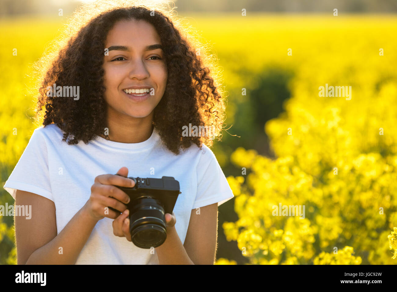 Beautiful happy mixed race African American girl teenager female young woman smiling outdoors in sunshine taking photographs with a camera Stock Photo