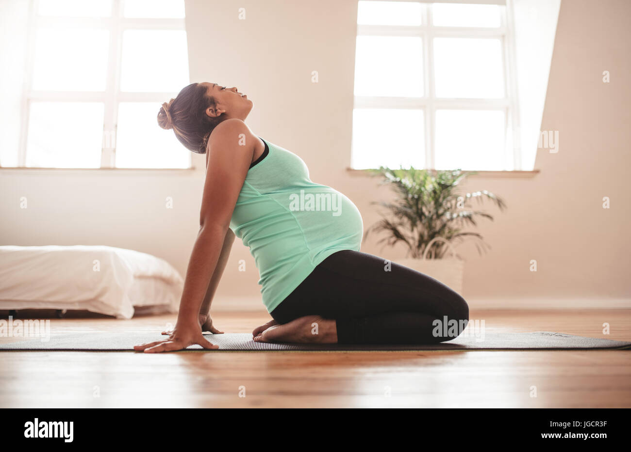 Side view of young pregnant woman kneeling on exercise mat and stretching backwards. Expectant mother practising pregnancy yoga at home. Stock Photo