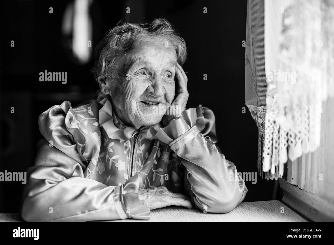 Elderly lady sits at the table and looking out the window. Black and white photo. Stock Photo