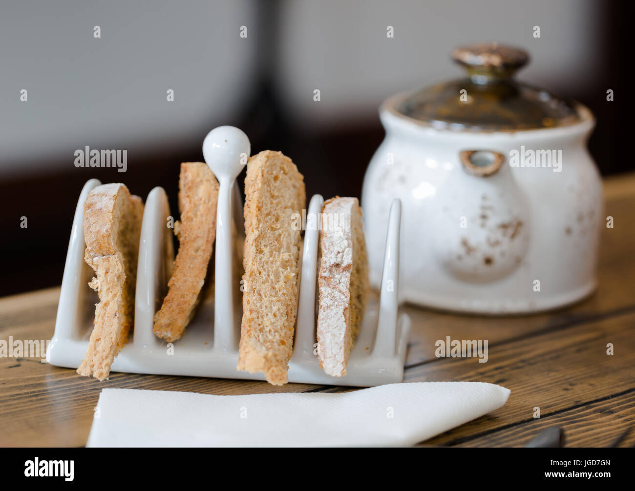 Four slices of toasted bread in toasts holder on wooden table close up. Foreground focus, tea pot in blurred background. Low angle close up view. Shal Stock Photo