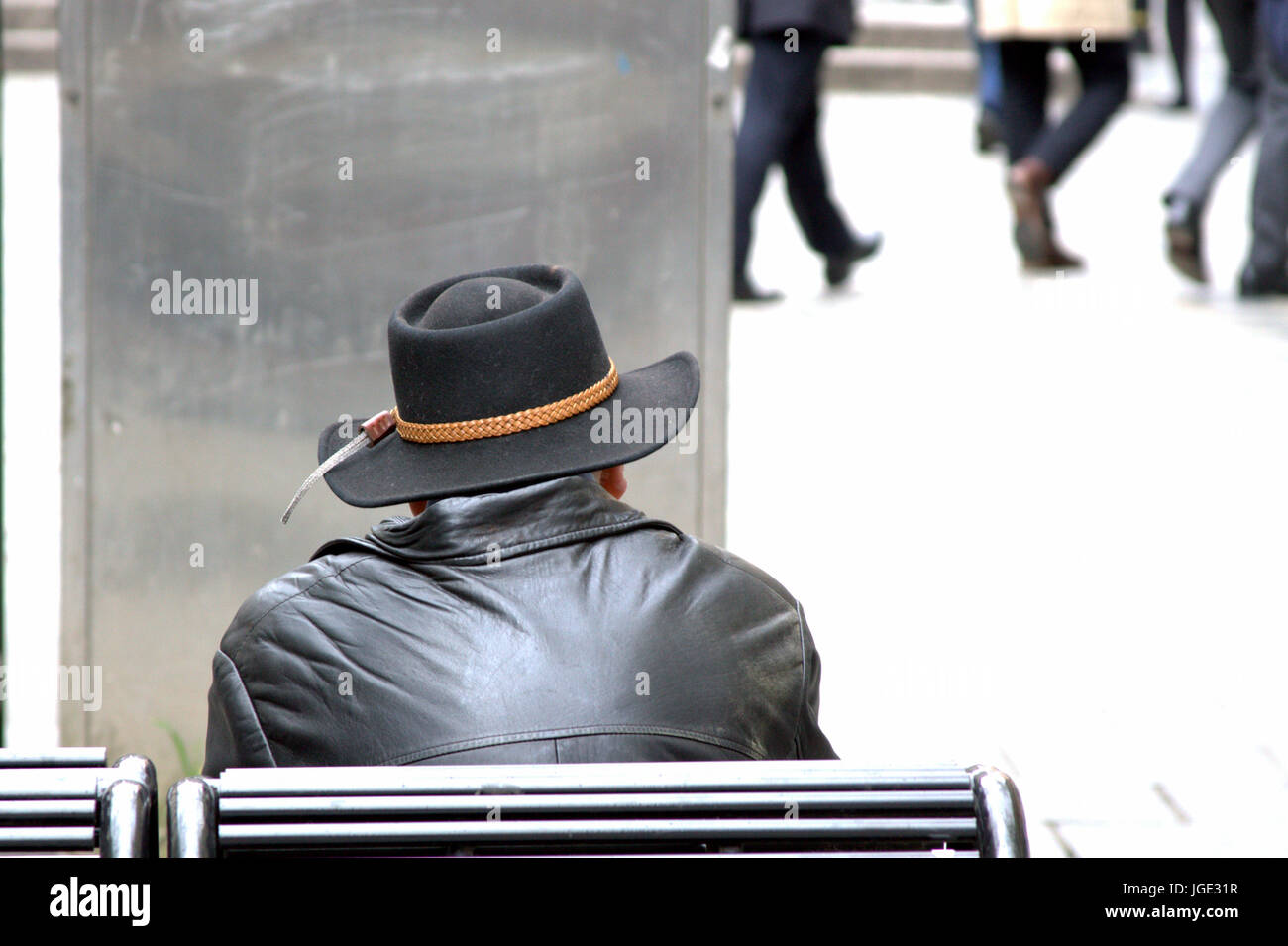 single man alone  sitting on a bench dressed in black with leather jacket and cowboy hat stetson  looking at people and the world going by Stock Photo