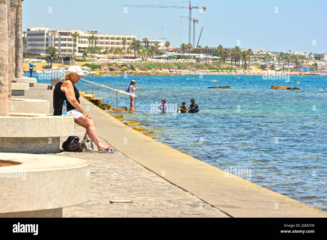 woman sitting on pafos harbour side enjoying the sunshine with scuba divers in back ground Stock Photo