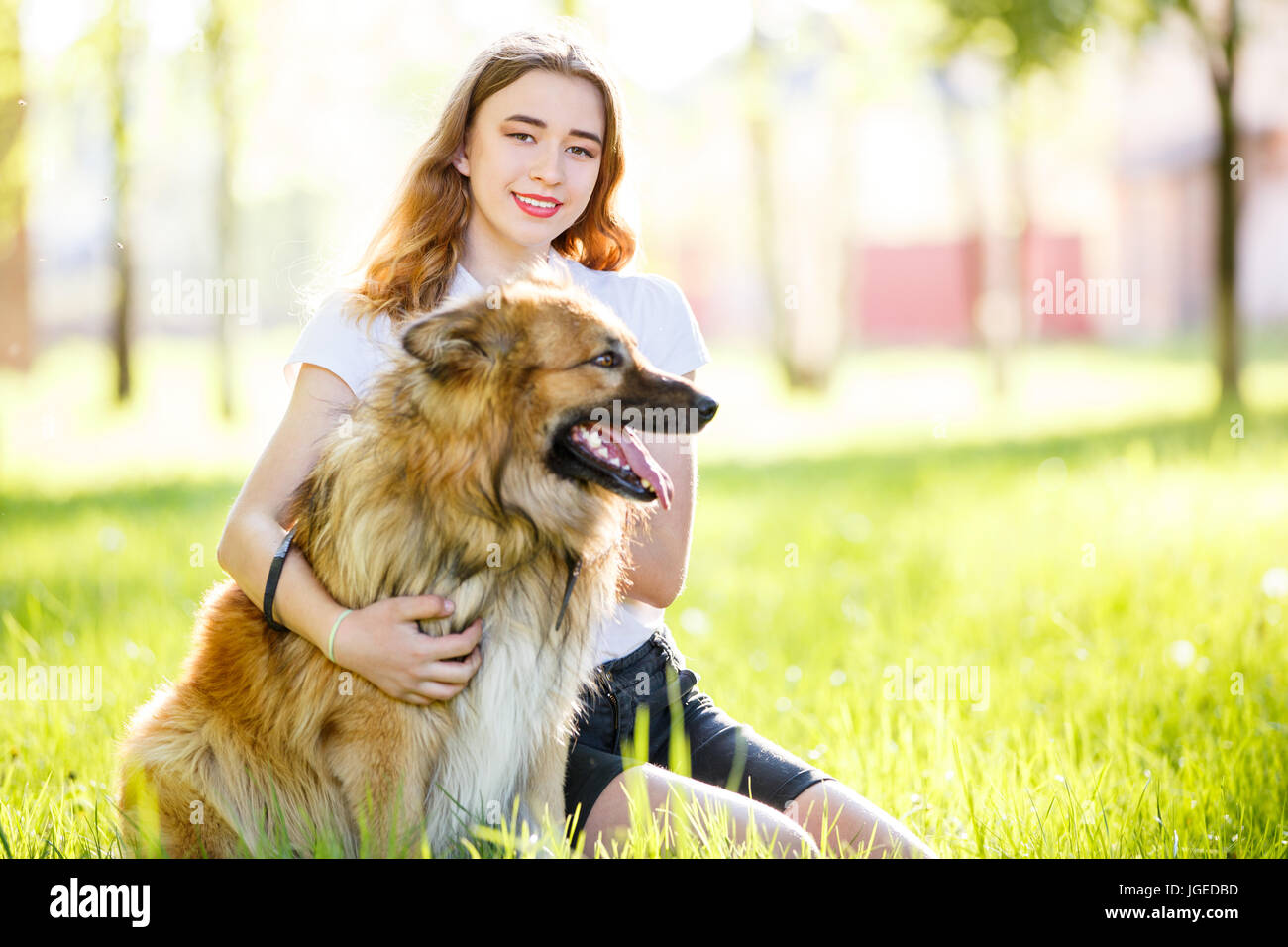 Teenage smiling girl in white shirt with her dog sitting on green grass ...
