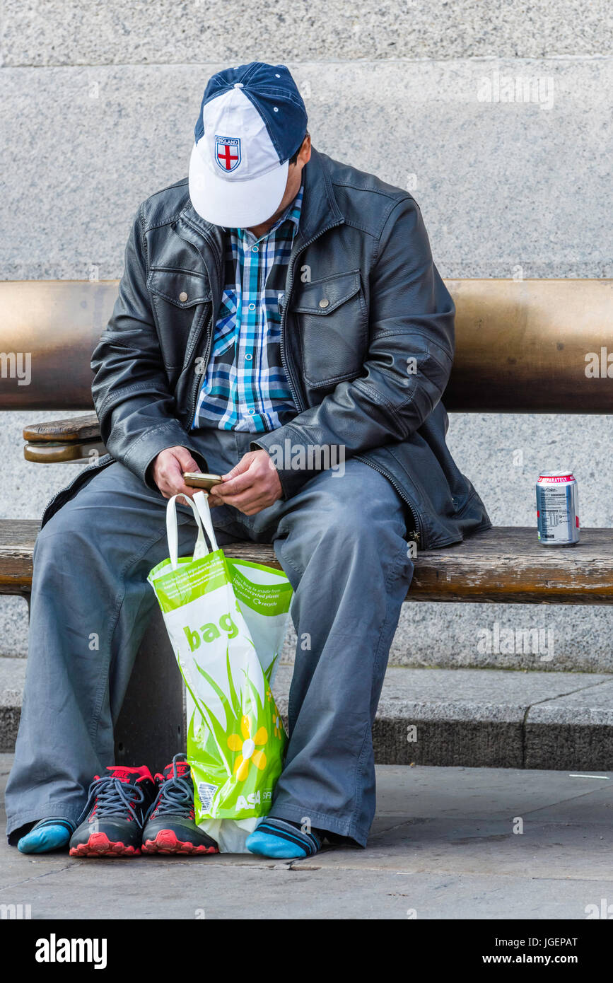 A middle aged man sitting on a bench with his shoes off, next to a can of drink, looking at his mobile phone/cell phone. Stock Photo