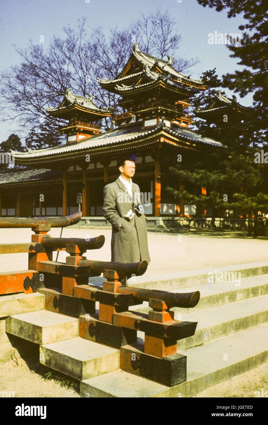 A tourist wearing a Kodak Brownie camera and an additional camera on straps around his neck walks past an ornate Japanese temple, Japan, 1955. Stock Photo