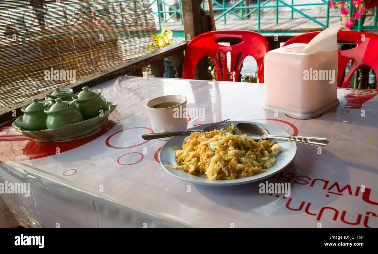 Plate of pad thai with spoon and fork on table next to condiment set and cup of tea at riverside restaurant. Bangkok, Thailand Stock Photo